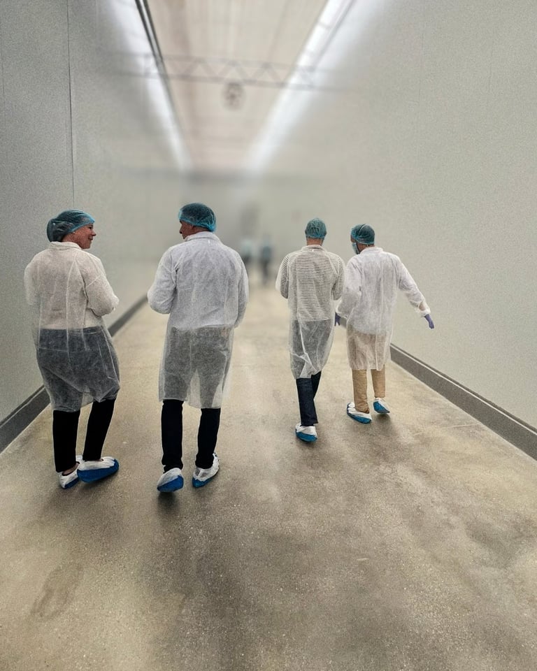 Four people wearing protective white lab coats and blue shoe covers walk through a sterile corridor with fluorescent lighting